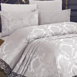 Könnte beinhalten: Ein luxuriöses Bettwäsche-Set mit einem hellgrauen Damastmuster. Die Bettwäsche umfasst einen Bettbezug, Kissenbezüge und Kissen. Der Bettbezug und die Kissenbezüge haben eine schwarze Spitzenborte. Das Bett steht vor einer dunkelblauen Wand.