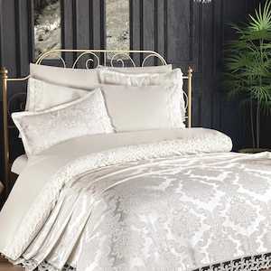 Puede incluir: Juego de cama color marfil con un estampado de damasco. El conjunto incluye un edredón, almohadas y fundas de almohada, todos con intrincados detalles de encaje. La estructura de la cama es dorada, sobre un fondo de paneles de madera oscura.