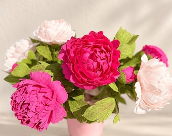 May include: A bouquet of handcrafted crepe paper peonies in shades of vibrant fuchsia and soft pale pink, featuring textured green paper leaves, arranged in a small, solid light pink container.