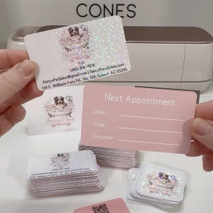 May include: Rectangular business cards with a holographic finish featuring a dog in a bathtub graphic, the text Fancy Paws, contact details, and an address. The reverse side is solid light pink with the text Next Appointment, Date, Time, and Groomer.