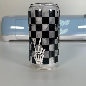 May include: A clear glass can with a black and white checkered pattern and a white skeleton hand making a peace sign.