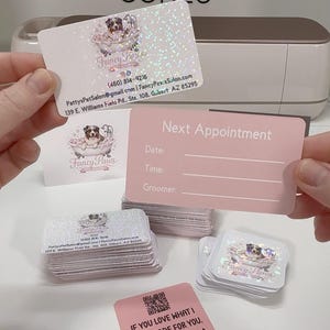 May include: A collection of rectangular and square business cards with holographic finishes. One card displays the Fancy Paws logo, contact details, and address. Another is a pink Next Appointment card. A square card features a QR code and review request.