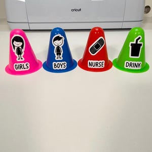 May include: Four plastic cones in pink, blue, red, and green. Each cone features a white sticker with a black graphic and text. The pink cone shows a girl and says GIRLS, the blue cone shows a boy and says BOYS, the red cone shows a bandage and says NURSE, and the green cone shows a drink cup and says DRINK.