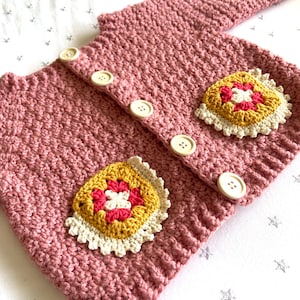 Puede incluir: Un cárdigan de punto rosa con botones blancos y dos aplicaciones de flores de ganchillo de colores.