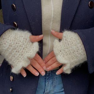 Peut inclure: Des mitaines en tricot sans doigts de couleur crème sont portées sur un pull crème. Un blazer bleu marine avec des boutons dorés et un jean complètent la tenue. Les gants semblent doux et texturés.