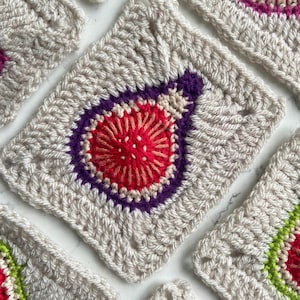 May include: Close-up of a crocheted square with a red, pink, and purple flower design in the center. The square is made with a beige yarn.