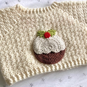 以下が含まれることがあります： 白の編み物で作られたベビーセーター。茶色、白、緑の編み物で作られたクリスマスプディングのアップリケが付いている。