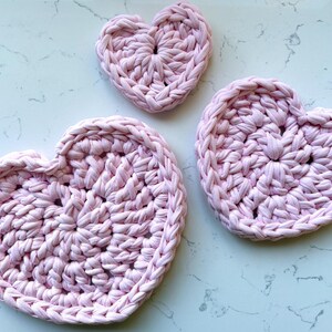 May include: Three light pink, heart-shaped crochet coasters. The coasters are made from a thick, textured yarn, with a visible stitch pattern. The hearts vary in size, with the largest measuring approximately 15 cm across.