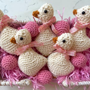 May include: A woven basket filled with handmade crochet geese and pink yarn. The geese are cream-colored with orange beaks and black eyes, each adorned with a pink gingham ribbon. Pink crochet balls are also in the basket.