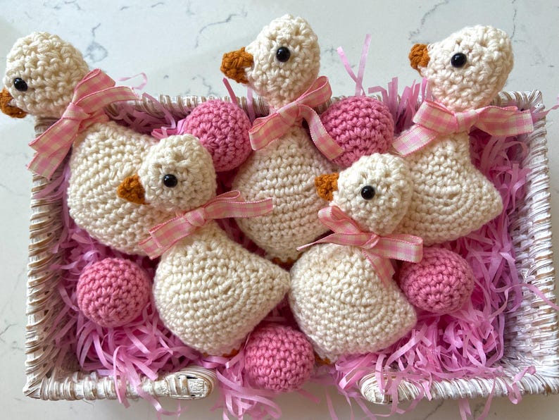 May include: A woven basket filled with handmade crochet geese and pink yarn. The geese are cream-colored with orange beaks and black eyes, each adorned with a pink gingham ribbon. Pink crochet balls are also in the basket.
