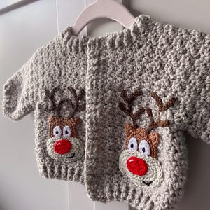 Puede incluir: Un cárdigan de punto de ganchillo gris con dos aplicaciones de reno de nariz roja. El cárdigan tiene cierre de botones y está hecho con un hilo suave y texturizado.