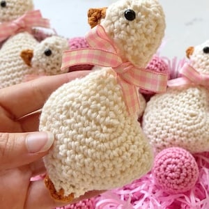 May include: Crocheted white duckling with a pink and white checkered ribbon bow. The duckling has a brown beak and black eyes. Other crocheted ducklings and pink yarn balls are in a white basket.