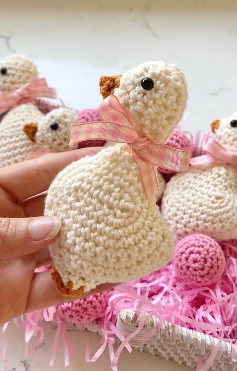 May include: Crocheted white duckling with a pink and white checkered ribbon bow. The duckling has a brown beak and black eyes. Other crocheted ducklings and pink yarn balls are in a white basket.
