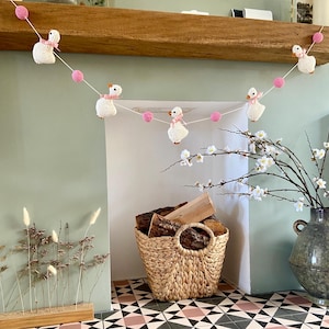 May include: A handmade garland featuring crocheted white geese with pink bows and pink yarn balls. The garland is strung across a wooden mantel. A woven basket holds firewood, and a vase holds flowering branches.