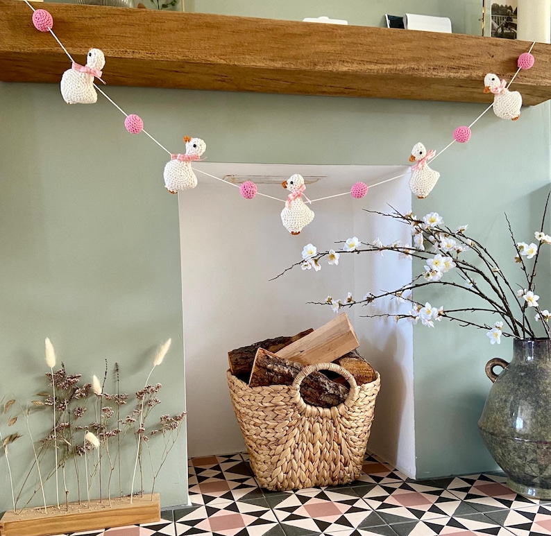 May include: A handmade garland featuring crocheted white geese with pink bows and pink yarn balls. The garland is strung across a wooden mantel. A woven basket holds firewood, and a vase holds flowering branches.
