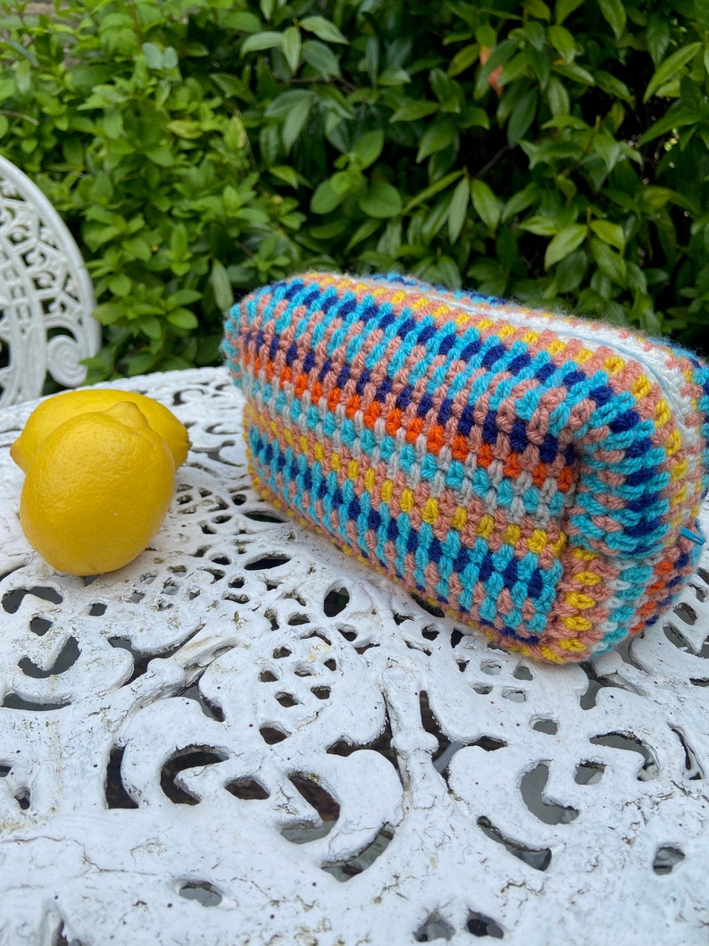 Może przedstawiać: Kolorowa, dziana torebka z paskami w kolorze niebieskim, r&oacute;żowym, ż&oacute;łtym, pomarańczowym i białym. Torebka leży na białym, ozdobnym stole.