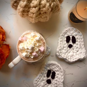 May include: A cozy autumn scene featuring a mug of hot chocolate topped with whipped cream and marshmallows, a lit candle, and three white crochet ghost coasters. A beige knitted pumpkin and orange roses complete the fall aesthetic.