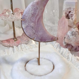 May include: A crescent moon-shaped crystal sculpture in shades of lavender and white, mounted on a bronze stand. The crystal sits on a white, fluffy base. Other crystals and decorative items are in the background.