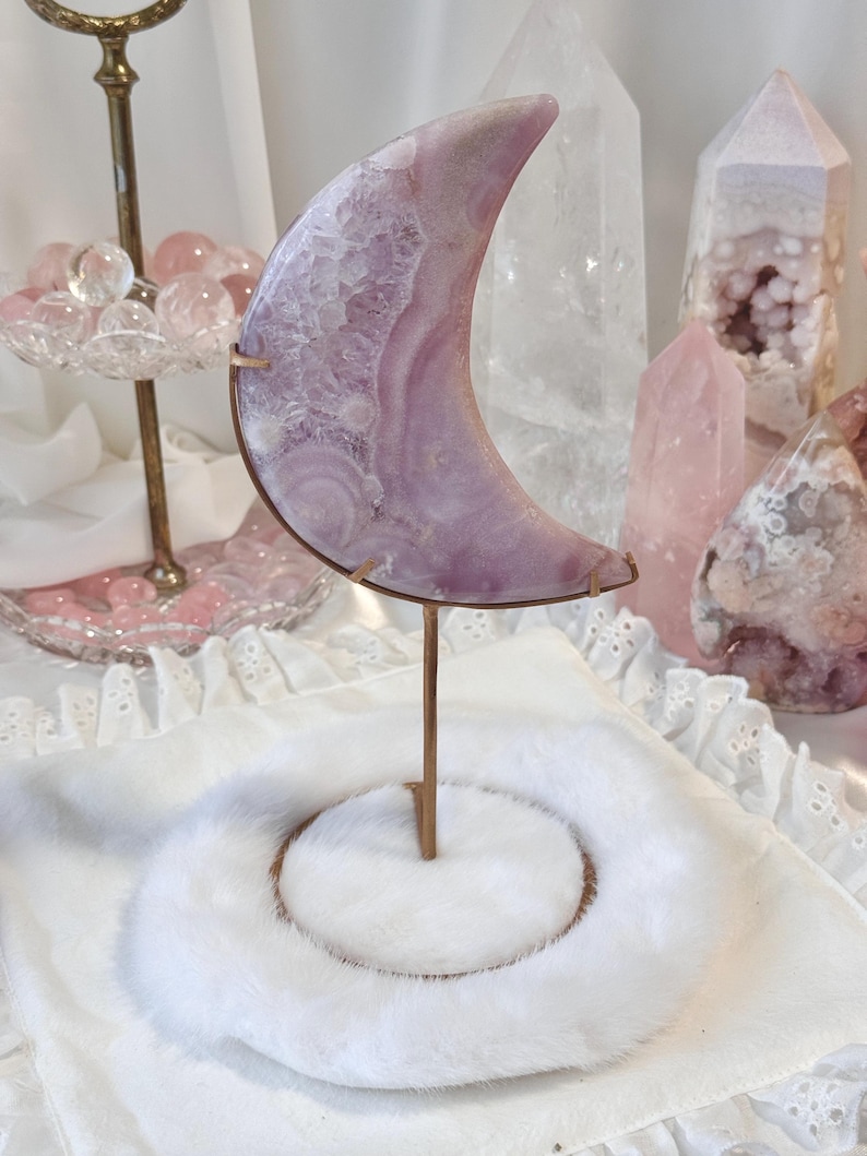 May include: A crescent moon-shaped crystal sculpture in shades of lavender and white, mounted on a bronze stand. The crystal sits on a white, fluffy base. Other crystals and decorative items are in the background.