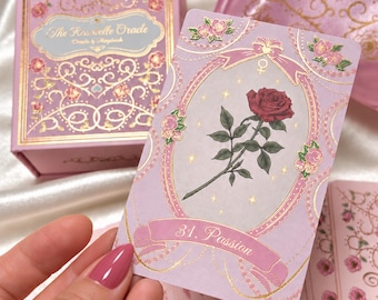 May include: A deck of oracle cards titled The Rosebelle Oracle, featuring a light pink box with gold foil floral accents and a matching satin drawstring bag. One card displays a red rose illustration, gold filigree, and the text 31. Passion.