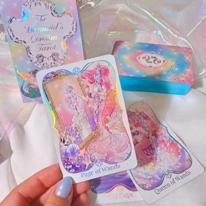 Könnte beinhalten: Das Tarot-Deck The Mermaid's Dream besteht aus einer Schachtel und Karten mit holografischen Akzenten. Die Karte Page of Wands zeigt eine Meerjungfrau, die in einen Spiegel mit einer Kerze blickt. Weitere Karten sind Queen of Wands und 5 of Cups.