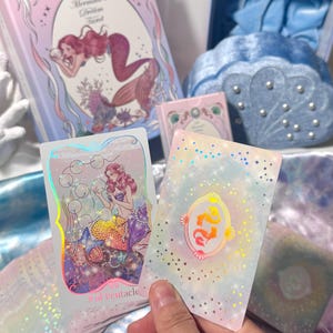 Może przedstawiać: Talia tarota zatytułowana The Mermaid's Dream Tarot, zawierająca jasnoniebieskie pudełko z ilustracją syreny, niebieski aksamitny woreczek w kształcie muszli z perłami oraz dwie karty przedstawiające syrenę z ośmioma pentaklami i gwiazdami.