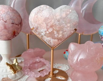 May include: A collection of pink quartz crystals and decorative items. The display includes a heart-shaped crystal on a gold stand, a sphere, crescent moons, a bracelet, and a cat figurine. The items are arranged on a pink surface.