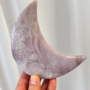 May include: A polished, crescent moon-shaped crystal in shades of lavender and white. The stone has a smooth, glossy finish with visible crystalline structures. The moon is held in a hand, showcasing its size and shape.