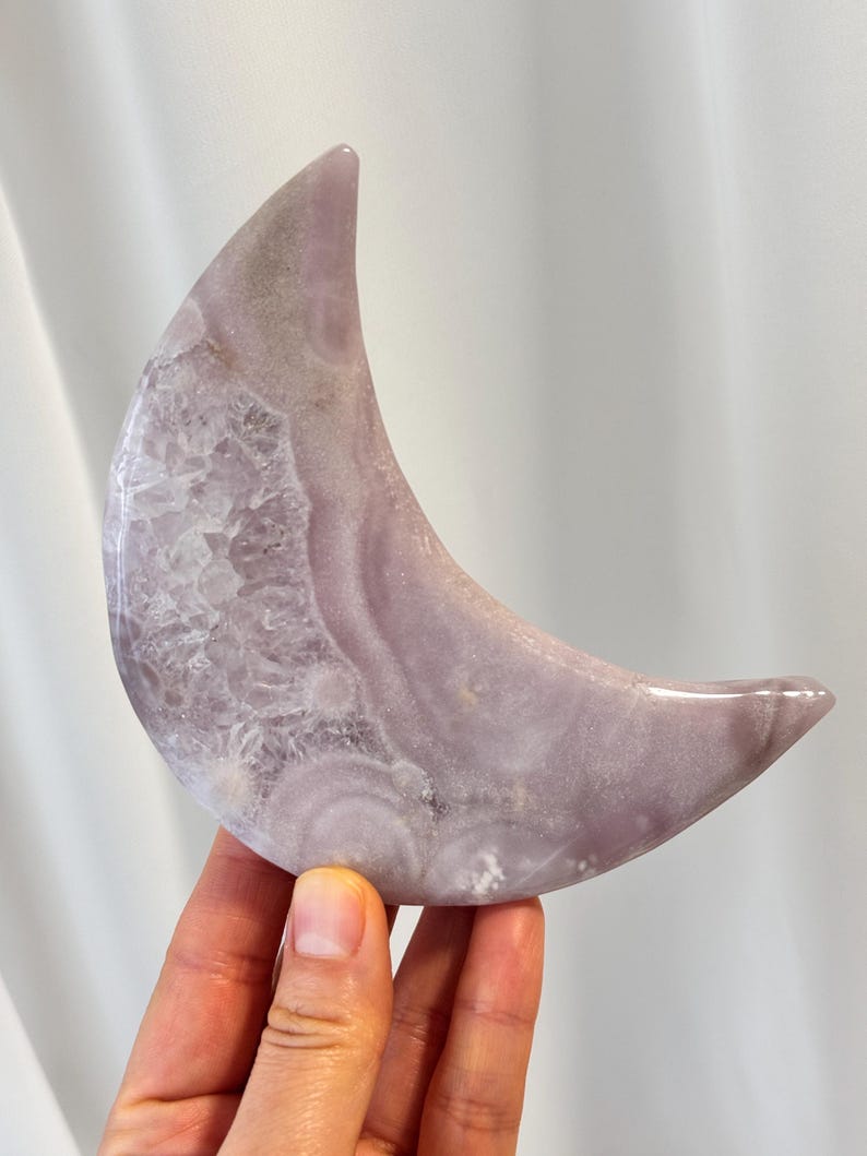 May include: A polished, crescent moon-shaped crystal in shades of lavender and white. The stone has a smooth, glossy finish with visible crystalline structures. The moon is held in a hand, showcasing its size and shape.