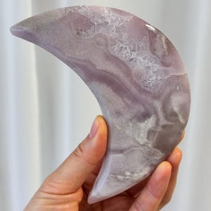 May include: A crescent moon-shaped decorative object. The object is a pale lavender color with white and gray marbling and crystalline inclusions. The smooth, polished surface reflects light. The object is held in a hand.