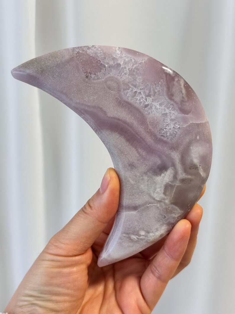 May include: A crescent moon-shaped decorative object. The object is a pale lavender color with white and gray marbling and crystalline inclusions. The smooth, polished surface reflects light. The object is held in a hand.
