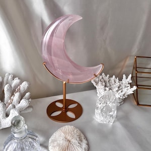Peut inclure: Une sculpture de lune en cristal rose sur un support en bronze. Le croissant de lune est en quartz rose translucide. Du corail décoratif et une bouteille en verre sont au premier plan. Le support a une base circulaire avec un motif floral.