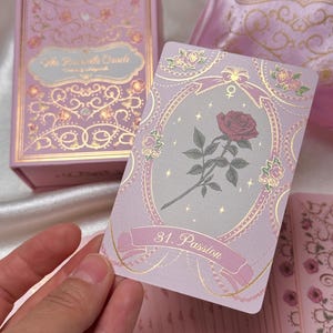 May include: A pink and gold tarot card with a red rose illustration. The card is labeled "31. Passion" and is part of "The Rosabelle Oracle" deck. The deck and box are pink with gold accents.