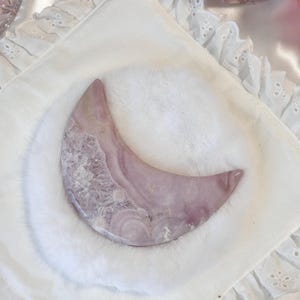 May include: A crescent-shaped, polished stone in shades of lavender and white, resting on a white, fluffy surface. The stone's surface displays a swirling pattern. The background includes a white, embroidered fabric and other pink objects.