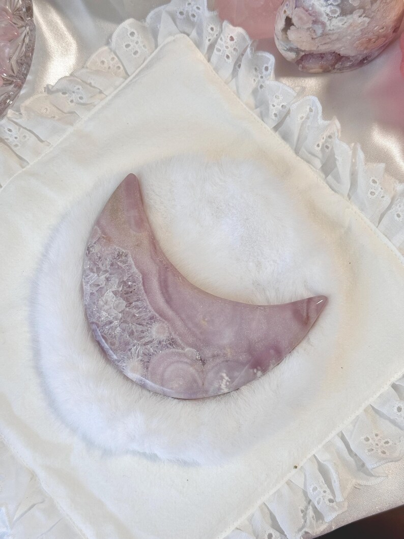 May include: A crescent-shaped, polished stone in shades of lavender and white, resting on a white, fluffy surface. The stone's surface displays a swirling pattern. The background includes a white, embroidered fabric and other pink objects.