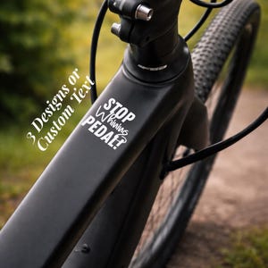 Pode incluir: Close-up de uma estrutura de bicicleta preta com o texto "STOP Whining PEDAL?" em branco. A imagem também inclui o texto "3 Designs or Custom Text". O pneu da bicicleta é visível no fundo.