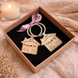 Pode incluir: Dois porta-chaves de madeira em forma de casa com argolas de metal, apresentados numa caixa de presente castanha com uma fita malva. Cada porta-chaves está gravado com o texto 2025, OUR FIRST HOME e os nomes Mailey e Jeff separados por um coração.