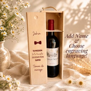 Peut inclure: Une boîte à vin en bois clair avec une poignée en corde contenant une bouteille de vin rouge. Le couvercle coulissant est gravé avec le prénom John, une icône de nœud papillon et le texte : TA MISSION Si tu l'acceptes DEVENIR MON SUPER Témoin de mariage.