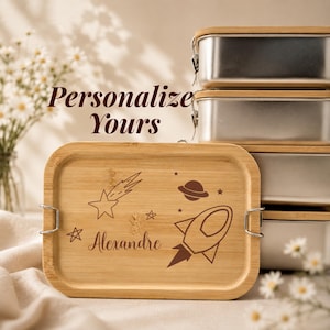 Può includere: Un portapranzo rettangolare in acciaio inossidabile con coperchio in bambù caratterizzato da un tema spaziale inciso al laser con un razzo, stelle, un pianeta e il nome Alexandre. Il testo Personalize Yours è visibile sopra i contenitori.