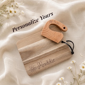 Peut inclure: Une planche à découper en bois clair et un outil à manche en bois assorti, tous deux gravés du prénom Maddie et d'une petite icône de fleur. Le texte Personalize Yours est visible au-dessus des objets, dotés d'une boucle en cuir noir.