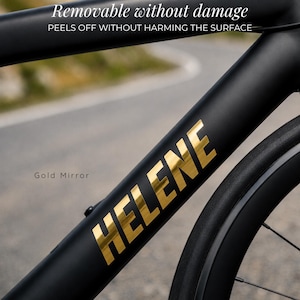 Pode incluir: Um adesivo com acabamento espelhado dourado com o texto HELENE aplicado em um quadro de bicicleta preto fosco. O texto acima diz Removable without damage e PEELS OFF WITHOUT HARMING THE SURFACE. O adesivo está no tubo diagonal do quadro.