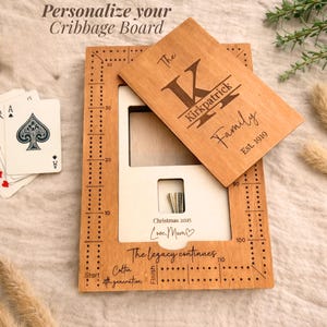 Op de afbeelding: Een houten cribbagebord met een gepersonaliseerde houten hoes. De hoes is gegraveerd met "The Kirkpatrick Family" en "Est. 1919". Het bord heeft een rechthoekig frame met gaten voor pinnen en de tekst "Christmas 2025 Love, Mom". Speelkaarten zijn zichtbaar.