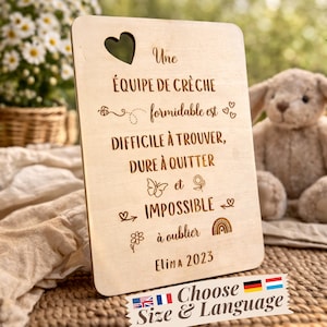 May include: A wooden plaque with a heart-shaped cutout. The plaque features engraved French text, including "Une Équipe de Crèche" and "Elina 2023". Decorative elements include hearts, a butterfly, a flower, and a rainbow. A banner at the bottom says "Choose Size & Language".