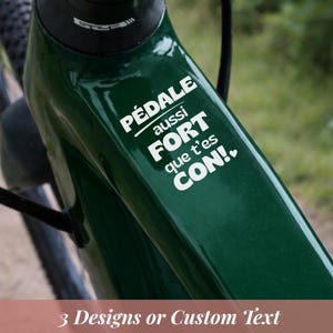 Peut inclure: Gros plan sur un cadre de vélo vert foncé avec le texte blanc "PÉDALE aussi FORT que t'es CON!". Le bas de l'image affiche le texte "3 Designs or Custom Text".