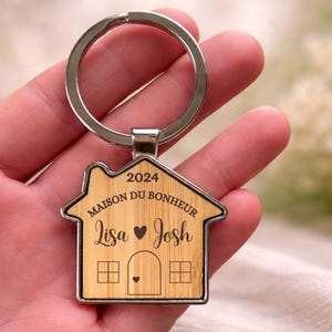 Peut inclure: Un porte-clés en forme de maison avec une incrustation en bois clair et un contour en métal argenté. Le bois est gravé avec le texte 2024, MAISON DU BONHEUR, les prénoms Lisa et Josh séparés par un cœur, et un dessin de maison.