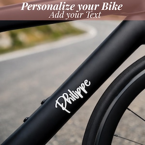 Peut inclure: Cadre de vélo noir avec le nom "Philippe" en écriture blanche. Le texte "Personnalisez votre vélo, Ajoutez votre texte" est en haut. La roue du vélo est visible sur le côté droit.