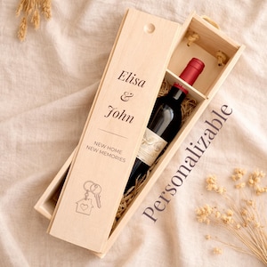 Peut inclure: Une boîte à vin en bois clair avec un couvercle coulissant gravé du texte Elisa & John, NEW HOME NEW MEMORIES, et d'une icône de maison et de clé. À l'intérieur, une bouteille de vin en verre foncé avec un bouchon rouge repose sur de la paille.