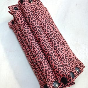 Könnte beinhalten: Gefalteter rosa Stoff mit schwarzem Leopardenmuster. Der Stoff ist mit schwarzen Druckknöpfen und Metallösen befestigt. Der Stoff ist fächerartig angeordnet.