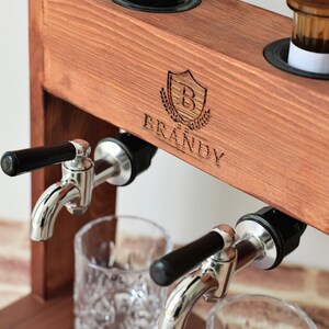 Może przedstawiać: Drewniany dozownik do alkoholu z dwoma kranami i dwoma szklankami. Dozownik ma etykietę z napisem "BRANDY" z tarczą i logo w kształcie litery "B".