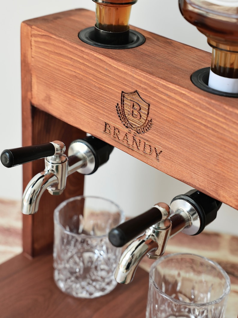 Może przedstawiać: Drewniany dozownik do alkoholu z dwoma kranami i dwoma szklankami. Dozownik ma etykietę z napisem "BRANDY" z tarczą i logo w kształcie litery "B".